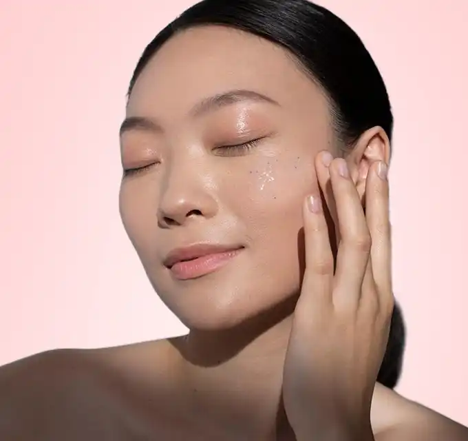 "Close-up of a woman with glowing, dewy skin gently touching her cheek on a soft pink background."