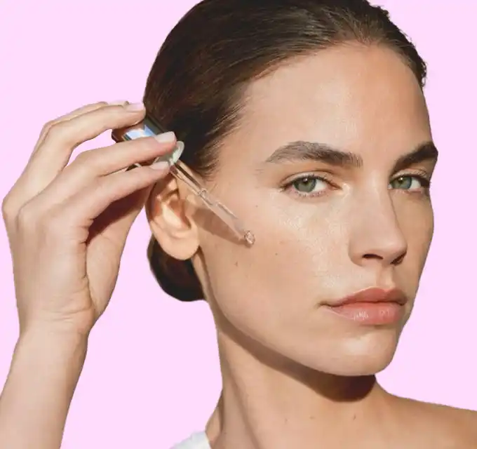 Clean portrait of a woman applying serum with a dropper on a pink background — neutral product/ingredient shot