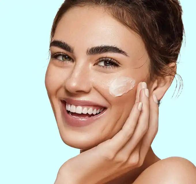 Smiling woman applying a dollop of cream to her cheek on a pale aqua background, showing hydrated, glowing skin