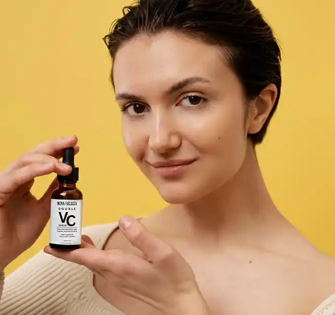 Close portrait of a woman presenting an amber VC serum bottle with both hands, skin appears even-toned and hydrated against a yellow backdrop