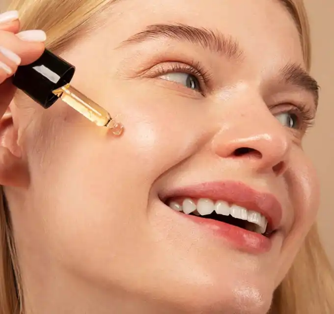 Close-up of a smiling fair-skinned woman as a golden serum drop falls from a glass dropper onto her cheek, skin looks dewy and radiant